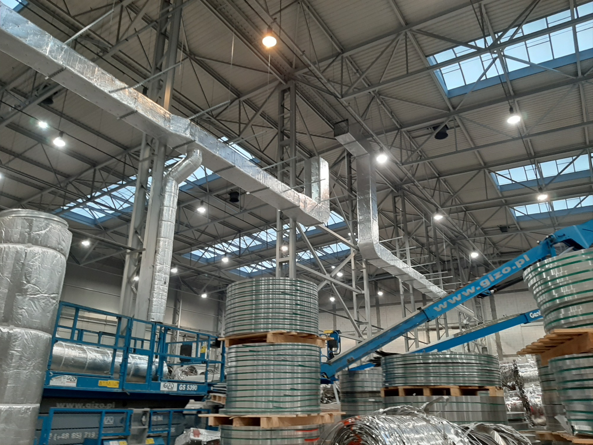 Wysokie hale magazynowe z widocznymi kanałami wentylacyjnymi pod sufitem, stosy blachy w kręgach na paletach i podnośnik koszowy z wysięgnikiem.