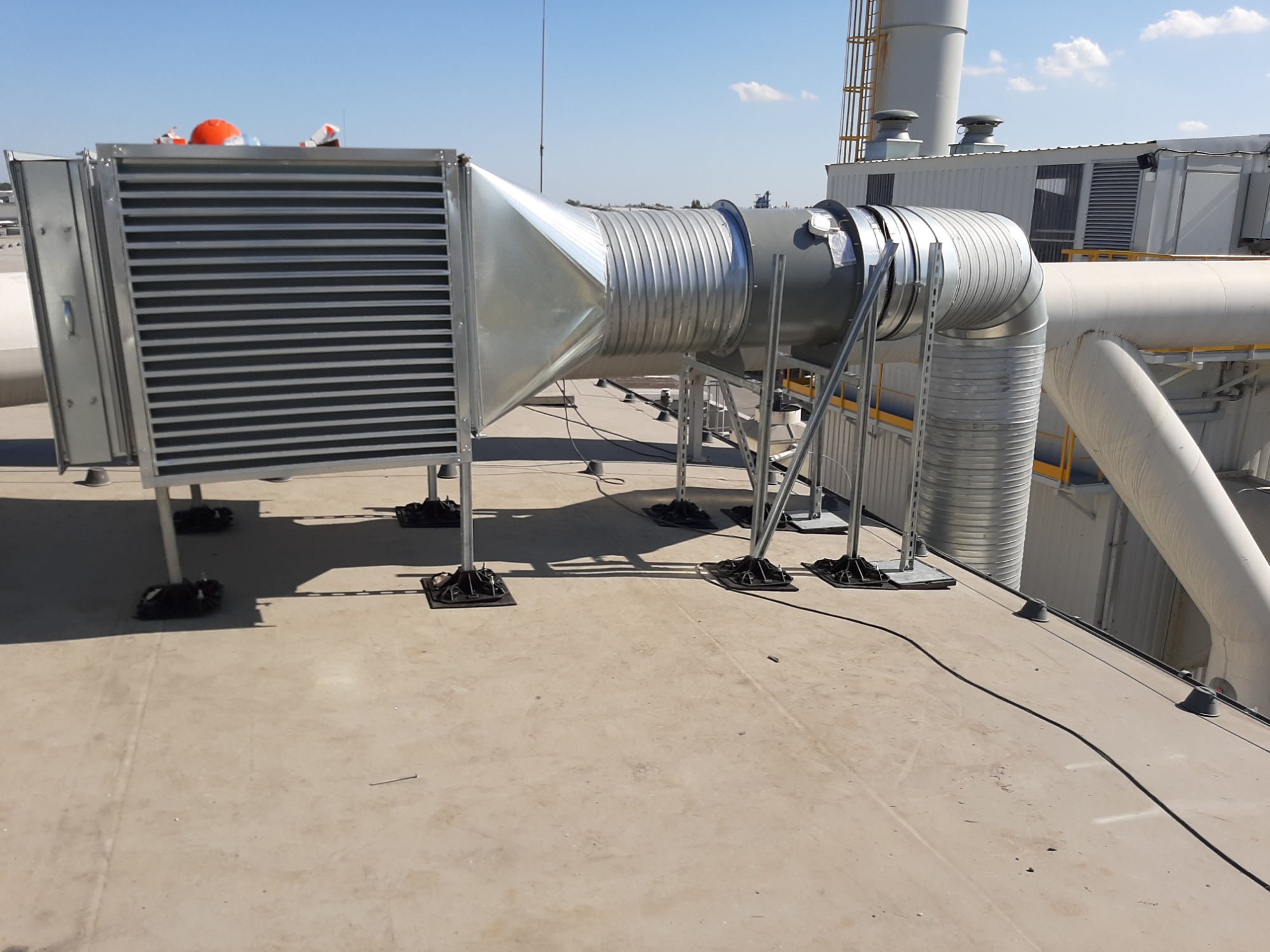 Instalacja wentylacji na dachu budynku komercyjnego z widocznymi kanałami wentylacyjnymi, tłumikiem i elementami mocującymi, z pomarańczową osłoną na górze