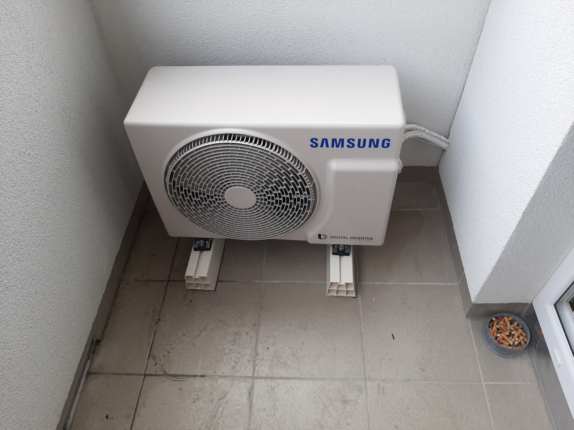 Zewnętrzna jednostka klimatyzacji Samsung Digital Inverter, zamontowana na wspornikach z tworzywa, z widocznymi kablami i popielniczką pełną niedopałków w rogu balkonu.