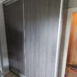 Szafa wnękowa z przesuwnymi drzwiami, wykończona w odcieniach szarości z widoczną strukturą drewna, zamontowana w pokoju z jasną podłogą.