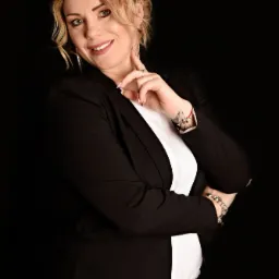 Portret uśmiechniętej blondynki w czarnej marynarce i białej koszuli na czarnym tle, z pierścionkiem i bransoletkami na rękach, pozującej z ręką pod brodą.