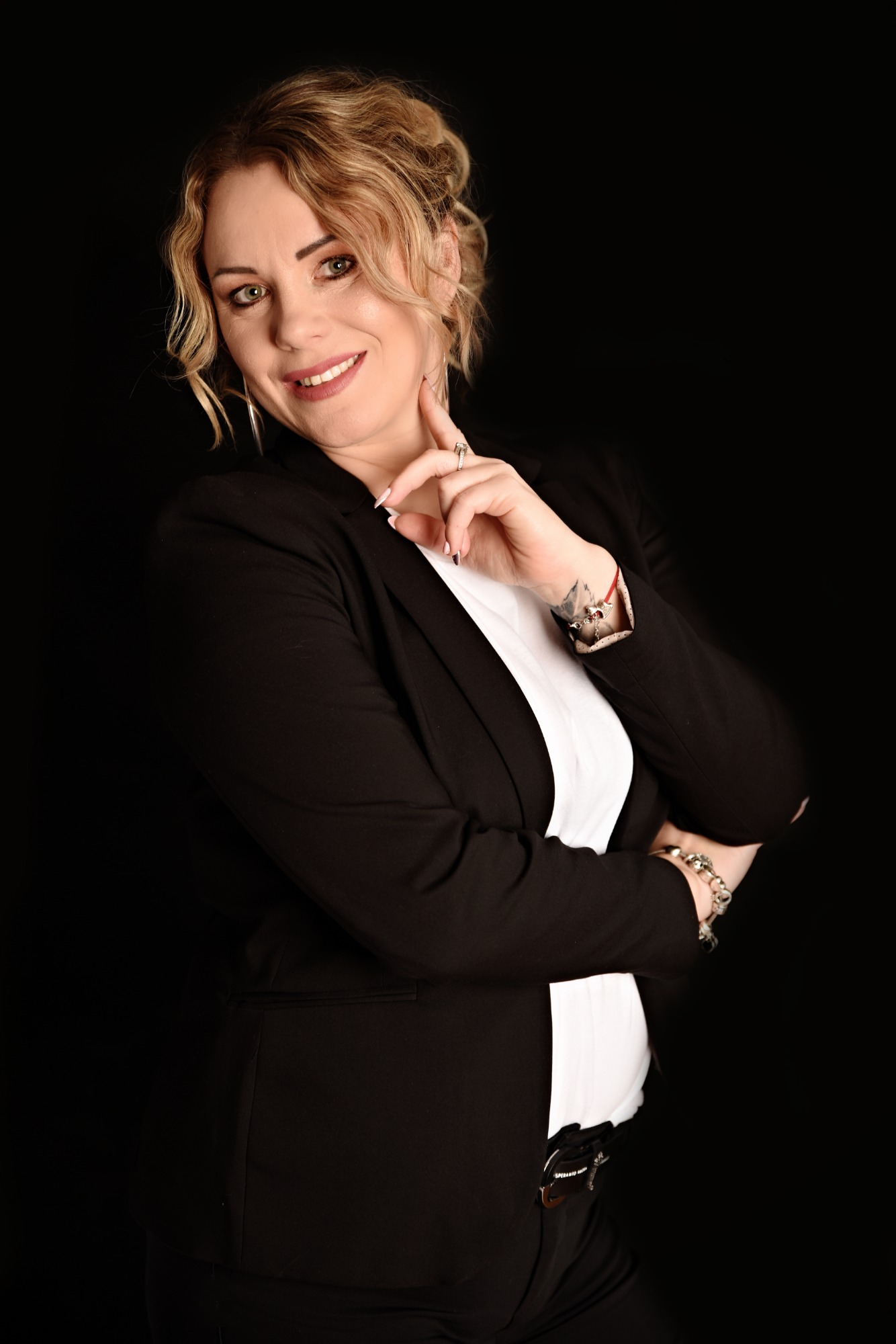 Portret uśmiechniętej blondynki w czarnej marynarce i białej koszuli na czarnym tle, z pierścionkiem i bransoletkami na rękach, pozującej z ręką pod brodą.