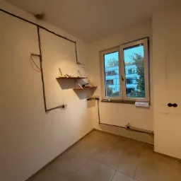 Wnętrze pokoju w trakcie remontu, widoczne przewody elektryczne rozprowadzone po ścianach, przygotowane punkty pod gniazdka, półki z narzędziami i materiałami budowlanymi, okno z widokiem...