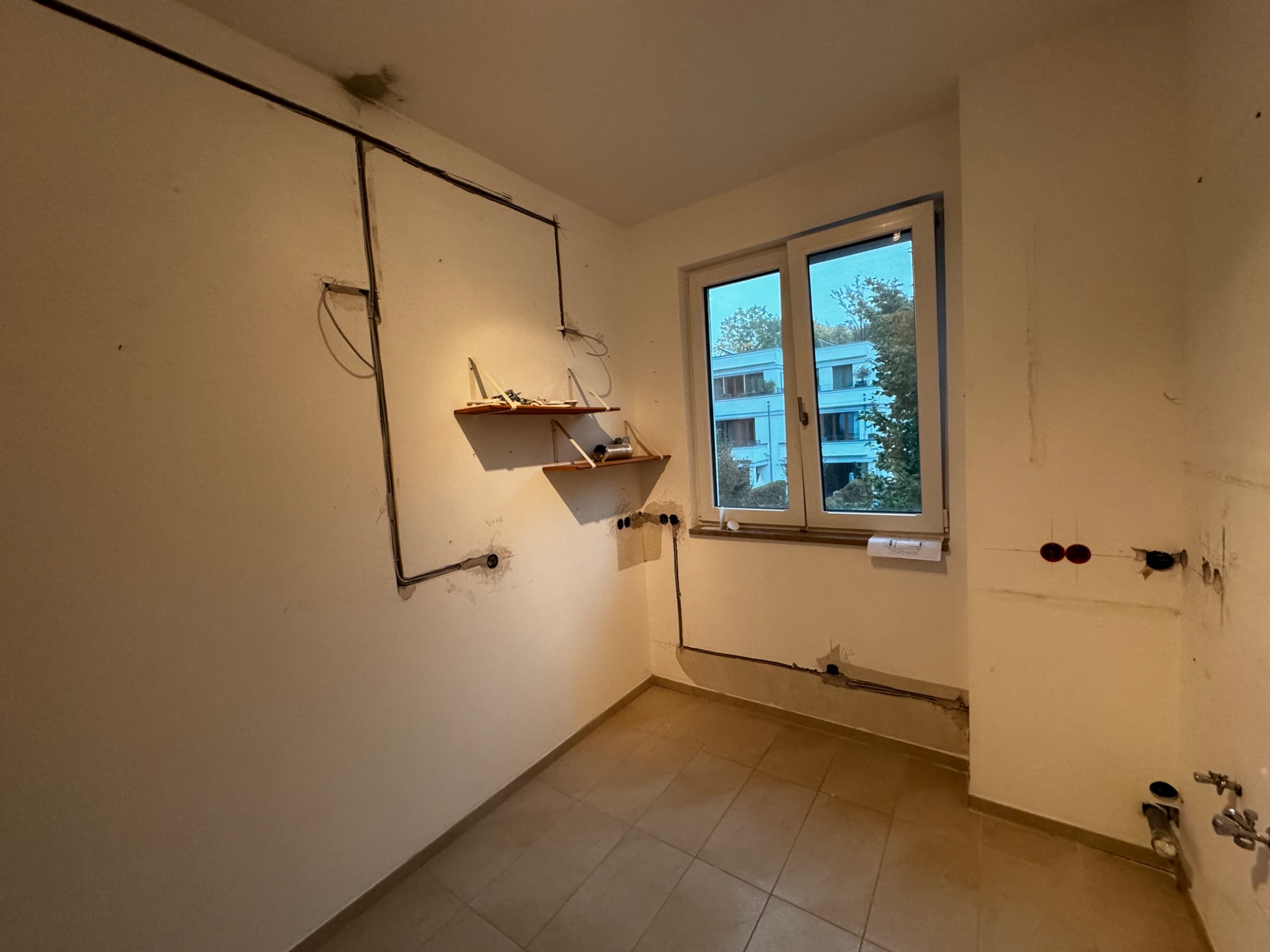 Wnętrze pokoju w trakcie remontu, widoczne przewody elektryczne rozprowadzone po ścianach, przygotowane punkty pod gniazdka, półki z narzędziami i materiałami budowlanymi, okno z widokiem...