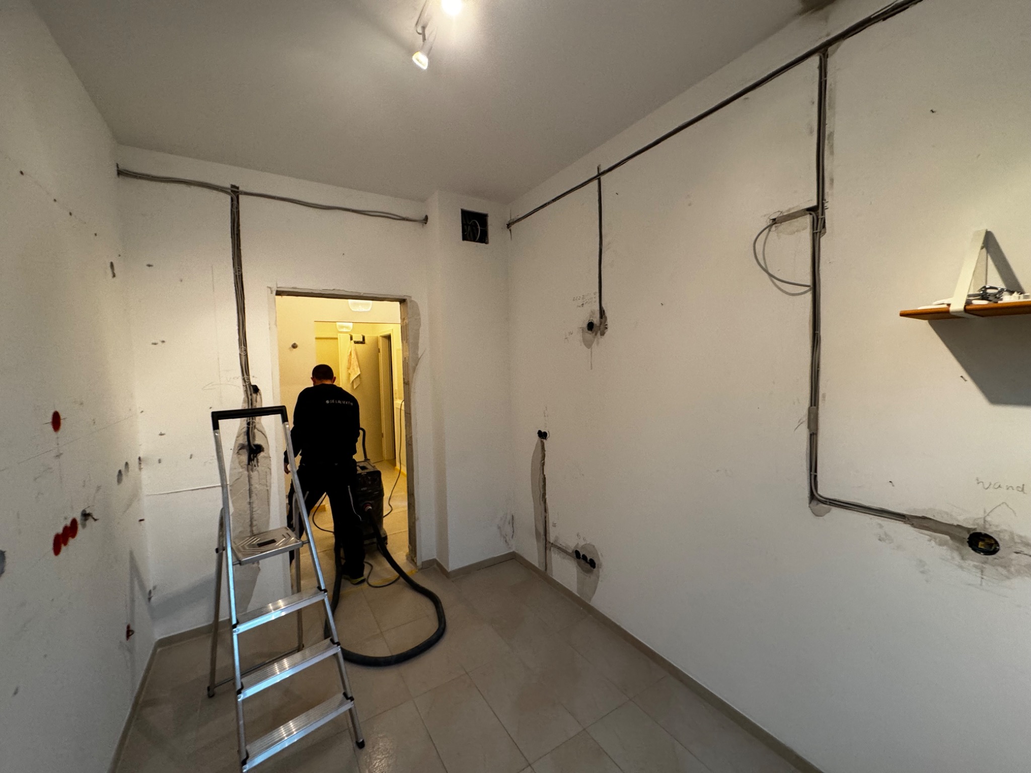 Wnętrze remontowanego pokoju z widocznymi przewodami elektrycznymi na ścianach, drabina aluminiowa i pracownik używający odkurzacza przemysłowego do usuwania pyłu.
