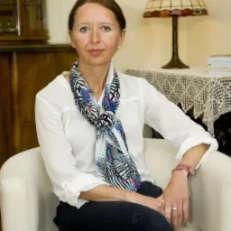 Portret kobiety, prawdopodobnie psycholog, siedzącej w białym fotelu w gabinecie z lampą Tiffany i dyplomami w tle, ubrana w białą koszulę i wzorzystą apaszkę.