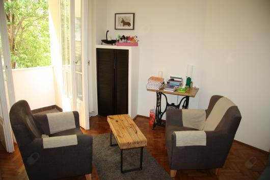 Jasny pokój z dwoma fotelami, drewnianym stolikiem kawowym i biurkiem ze starą maszyną do szycia w roli podstawy, na ścianie obrazek z koniem, widok na balkon.