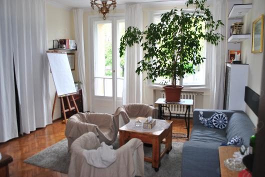 Jasne wnętrze gabinetu z trzema fotelami przykrytymi beżowymi narzutami, stolikiem kawowym, sofą z poduszkami i dużą rośliną doniczkową na tle okna.