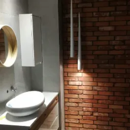 Nowoczesna łazienka z umywalką nablatową, szafką z szufladami, okrągłym lustrem z drewnianą ramą i oświetleniem punktowym oraz ścianą z czerwonej cegły.