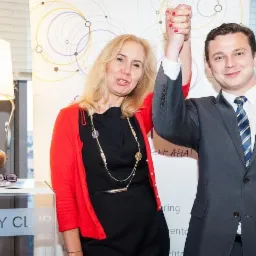 Kobieta w czerwonym kardiganie i mężczyzna w garniturze trzymają się za ręce na tle ściany z abstrakcyjnym wzorem kół. Na pierwszym planie stolik z lampą w kształcie dzbanka i okularami na otwartej...