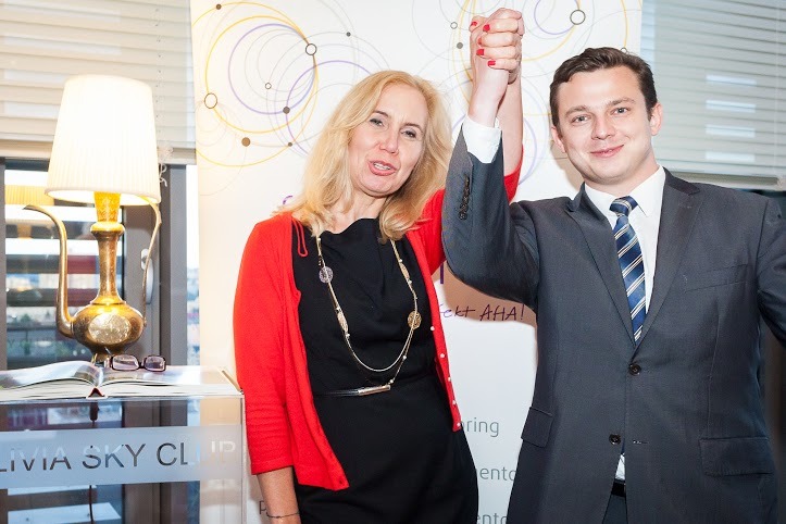 Kobieta w czerwonym kardiganie i mężczyzna w garniturze trzymają się za ręce na tle ściany z abstrakcyjnym wzorem kół. Na pierwszym planie stolik z lampą w kształcie dzbanka i okularami na otwartej...