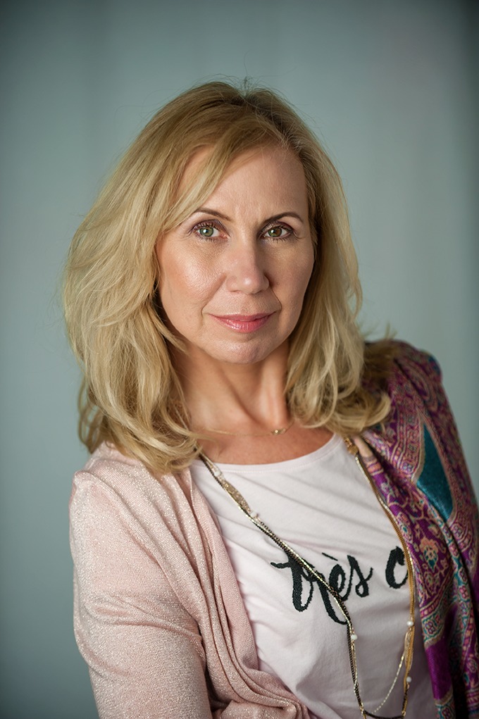 Portret kobiety o blond włosach, ubrana w różową bluzkę z napisem i wzorzystą chustę na ramieniu, patrząca prosto w obiektyw na neutralnym tle.