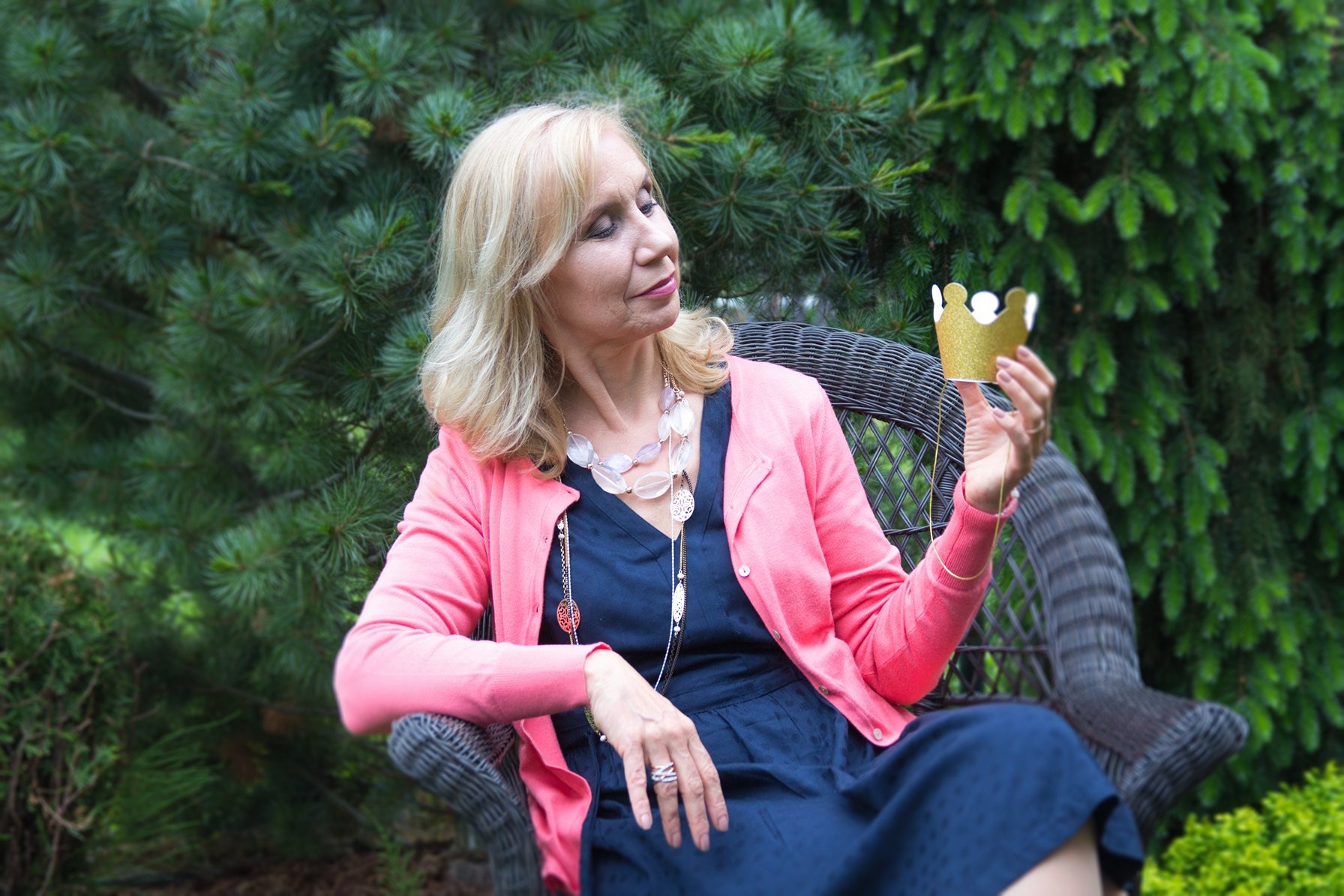 Kobieta siedzi w wiklinowym fotelu na tle zielonych krzewów, trzymając w dłoni złotą, papierową koronę.