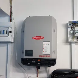 Szary inwerter Fronius Symo zamontowany na białej ścianie, obok otwartej skrzynki z bezpiecznikami i drugiej skrzynki elektrycznej z widocznymi przewodami.