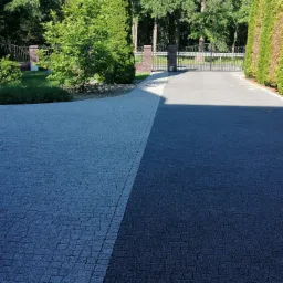 Dwukolorowy podjazd z kostki, jasnoszara i ciemnoszara, prowadzący do bramy wjazdowej z ceglanymi słupkami, otoczony zielenią drzew i krzewów.