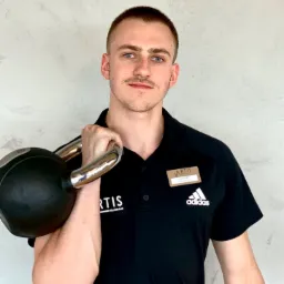 Młody mężczyzna z niebieskimi oczami, w czarnej koszulce polo z logo Artis i Adidas, trzyma na ramieniu czarny kettlebell ze srebrnym uchwytem, na tle jasnoszarej ściany.
