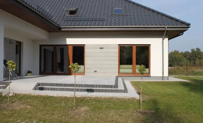 Nowoczesny dom jednorodzinny z szarą dachówką, białą elewacją z elementami szarego panelu, drewnianą stolarką okienną i brukowanym tarasem z niskimi schodami, otoczony trawnikiem z młodymi drzewkami.