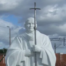 Biała marmurowa statua Jana Pawła II z metalowym krzyżem na tle pochmurnego nieba, ogrodzenia z drewnianymi sztachetami i linii kolejowej.