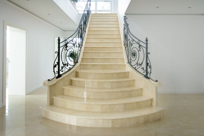 Marmurowe schody z kutą, ozdobną balustradą w jasnym, minimalistycznym wnętrzu. Widoczne detale wykończenia i elegancki design.