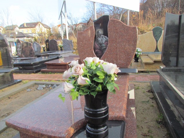 Pomnik z czerwonego granitu z wizerunkiem religijnym, ozdobiony bukietem sztucznych róż w czarnym wazonie, na tle innych nagrobków.