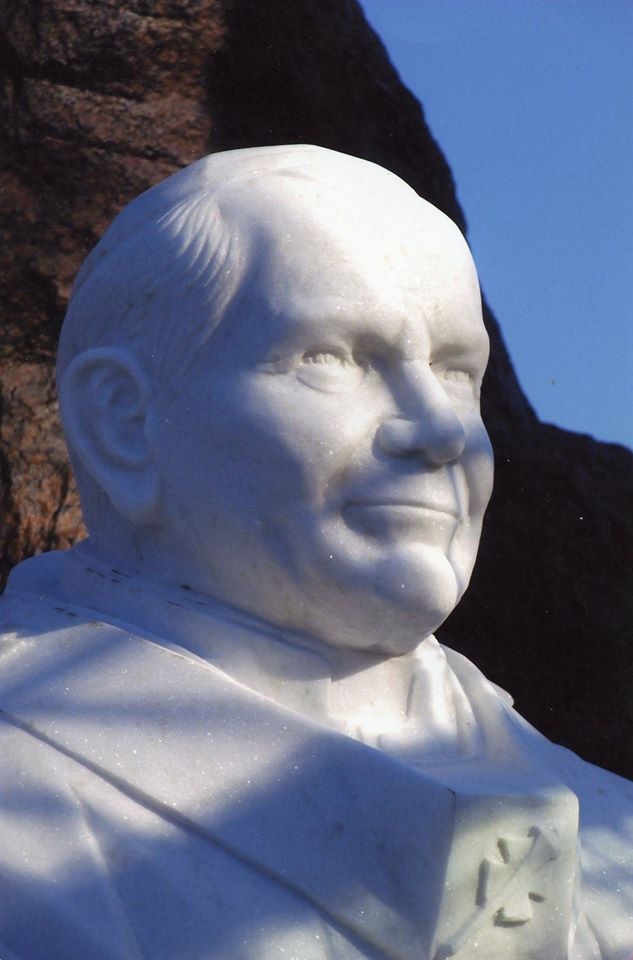 Biały marmurowy portret Jana Pawła II w plenerze, z widocznym fragmentem stroju papieskiego i tłem skalnym pod błękitnym niebem.