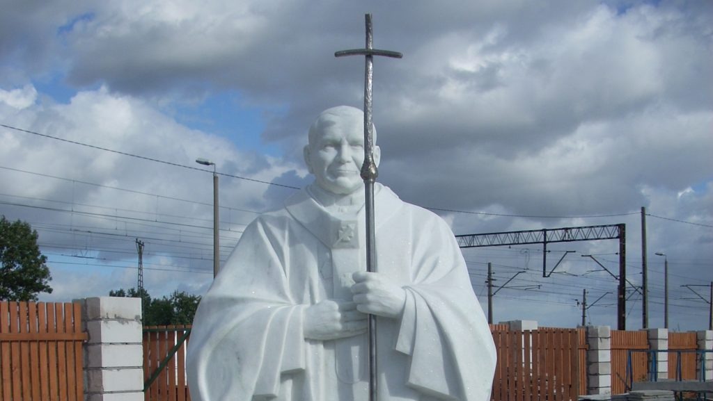 Biała marmurowa statua Jana Pawła II z metalowym krzyżem na tle pochmurnego nieba, ogrodzenia z drewnianymi sztachetami i linii kolejowej.