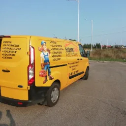 Żółty samochód dostawczy z grafiką hydraulika i informacjami o usługach elektrycznych, hydraulicznych, szklarskich, dekarskich i gazowych, zaparkowany na parkingu.