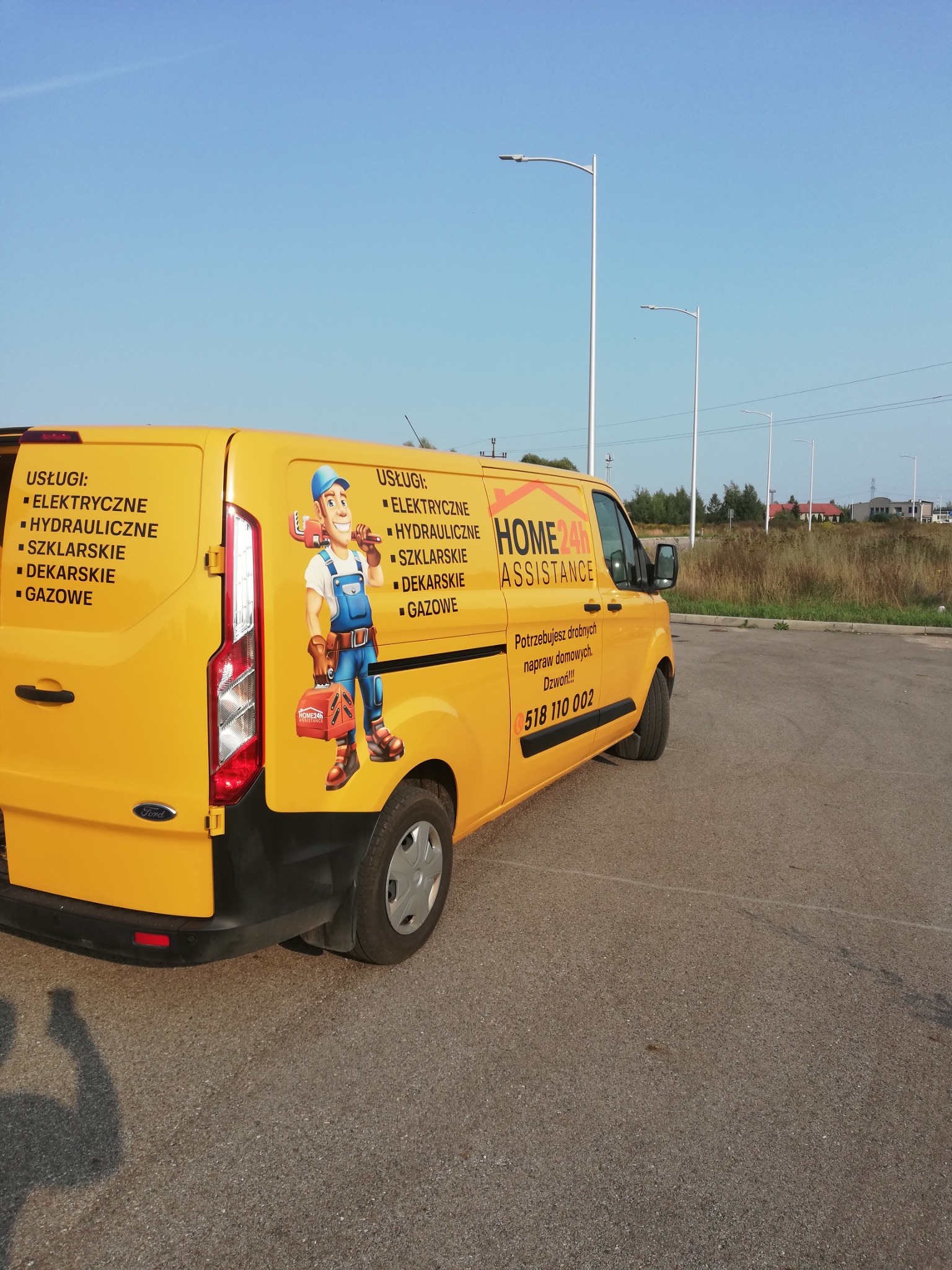 Żółty samochód dostawczy z grafiką hydraulika i informacjami o usługach elektrycznych, hydraulicznych, szklarskich, dekarskich i gazowych, zaparkowany na parkingu.