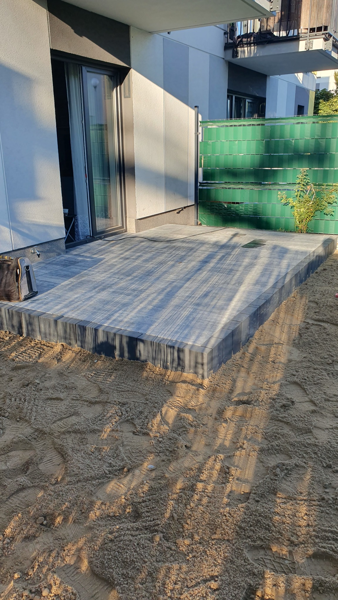 Nowo ułożony taras z szarych płyt betonowych, otoczony piaskiem i zieloną osłoną balkonową, widoczne ślady butów w piasku.