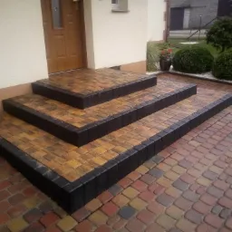 Trzystopniowe schody wejściowe wyłożone kostką brukową w odcieniach brązu i żółci, obrzeża z czarnej kostki, widok z góry na posesję z kostki brukowej, zieleń i elewacja budynku w tle.