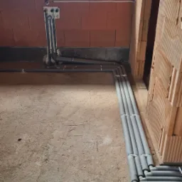 Szare rury instalacyjne w izolacji ułożone wzdłuż ściany z cegły ceramicznej dziurawki w surowym stanie, widoczne wyprowadzenia instalacji w ścianie i fragment okna z widokiem na zewnątrz.