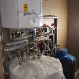 Nowa instalacja kotła grzewczego i zasobnika ciepłej wody, zabezpieczone folią ochronną, z widocznym filtrem wody i rurami w izolacji, na tle płytek ceramicznych.