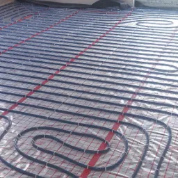 Rozłożone rury ogrzewania podłogowego na folii z czerwoną kratką, widoczne spinki mocujące, fragment okna w tle.