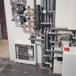 Szereg szarych, zaizolowanych rur instalacji grzewczej z manometrami i pompami obiegowymi, zamontowanych na białej ścianie w kotłowni na ciemnej, kwadratowej podłodze.