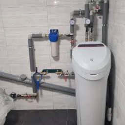 Biały zmiękczacz wody Viessmann z filtrem mechanicznym i manometrami, zamontowany na szarych rurach w pomieszczeniu z szaro-białymi płytkami.