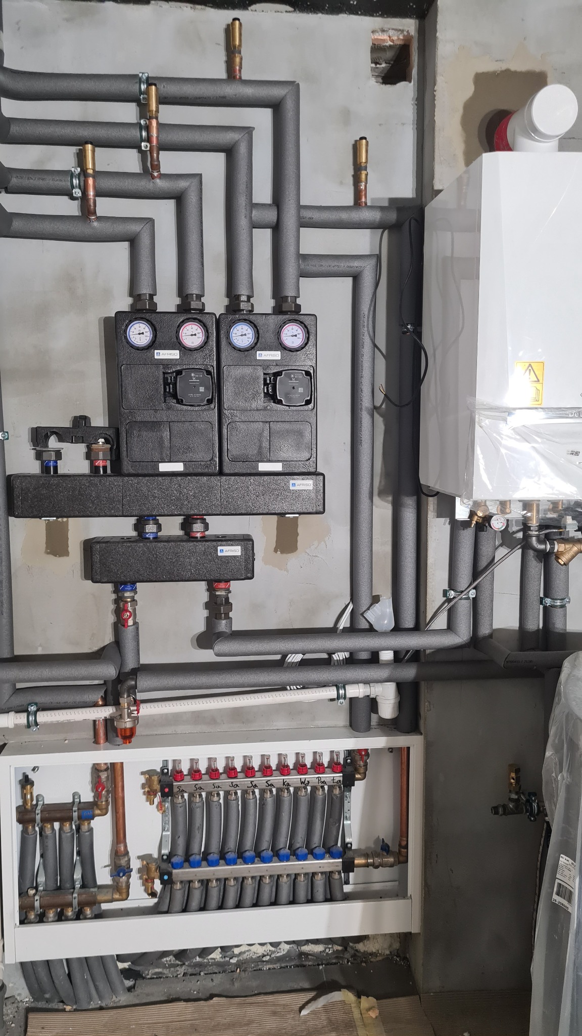 Rozdzielacz ogrzewania podłogowego z licznymi pętlami, zamontowany w szafce, z doprowadzonymi rurami w izolacji, obok wiszący piec, manometry na rozdzielaczach, surowa ściana w tle.