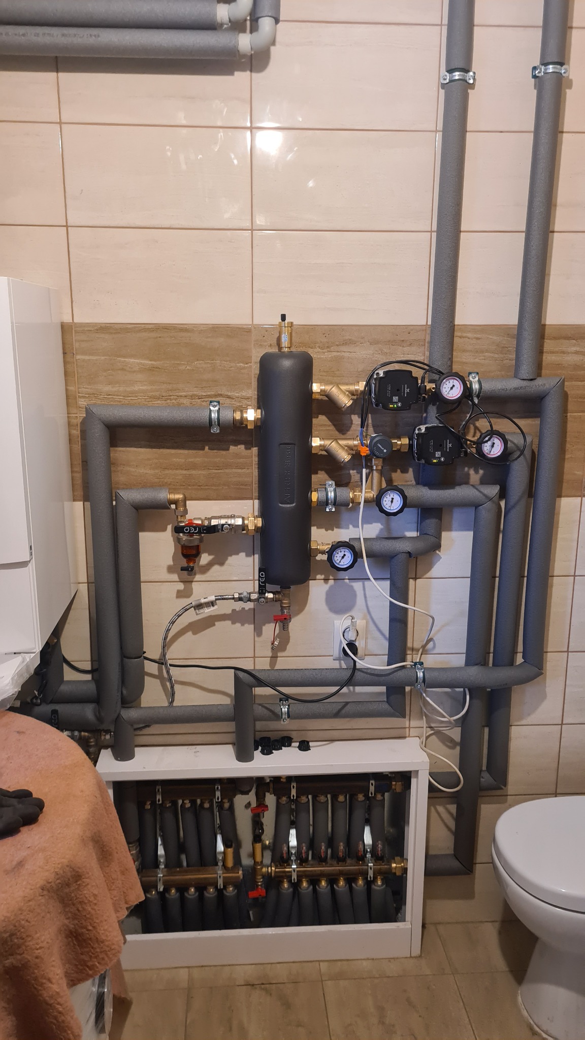 Rozdzielacz grzewczy z pompami obiegowymi, manometrami i rurami izolowanymi termicznie, zamontowany na ścianie z płytek ceramicznych w kotłowni.