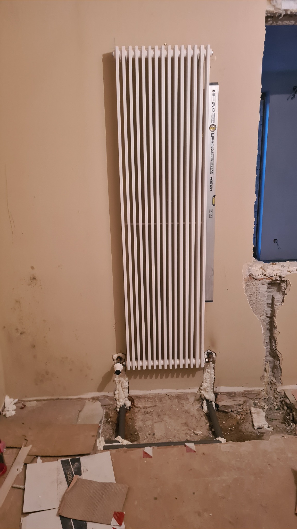 Montaż grzejnika panelowego z poziomnicą, widoczne rury instalacji grzewczej osadzone w pianie montażowej, ślady wilgoci na ścianie i wykończenie ściany obok otworu okiennego w trakcie prac...