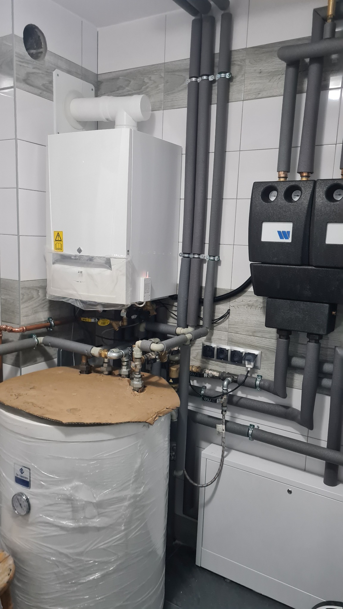 Biały piec centralnego ogrzewania z podłączonymi rurami w izolacji, zbiornik wody owinięty folią ochronną, widoczne elementy instalacji i gniazdka elektryczne na tle białych płytek.