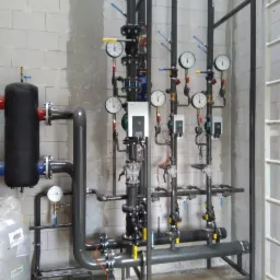 Złożona instalacja rur i zaworów z manometrami w pomieszczeniu z surowymi ścianami z bloków betonowych, widoczne elementy spawane.