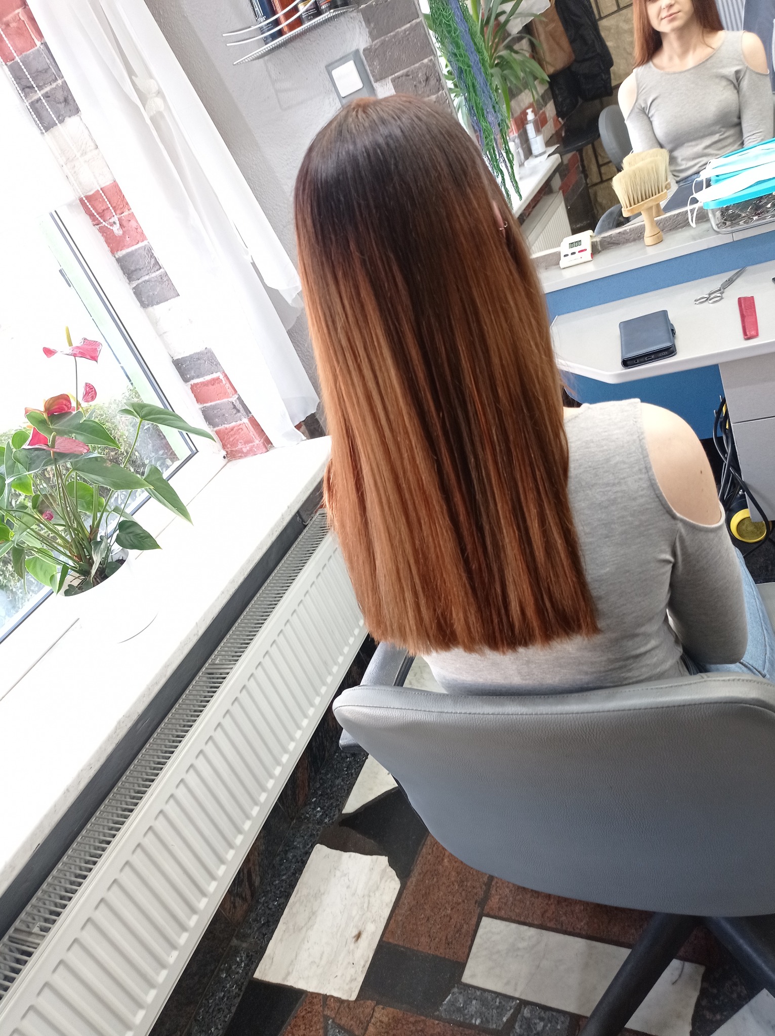 Kobieta siedząca w fotelu fryzjerskim, prezentująca świeżo obcięte, proste włosy w odcieniach brązu, z widocznym przejściem kolorów od ciemnego u nasady po jaśniejszy na końcach. W tle lustro, okno...