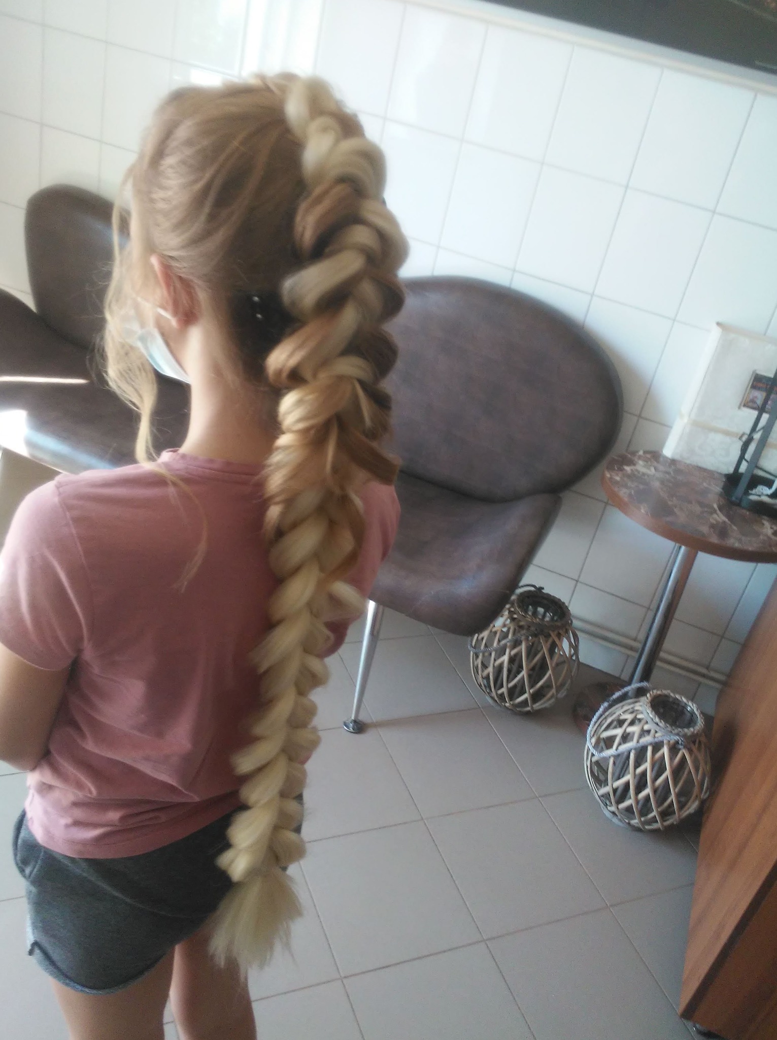 Dziewczynka z długim, grubym warkoczem ombre, przechodzącym od blondu do brązu, widziana od tyłu, w tle krzesła i dekoracyjne lampiony.