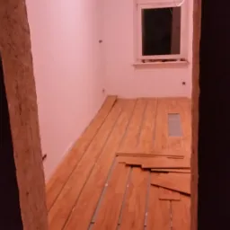 Układanie paneli podłogowych w pokoju z widoczną starą futryną i oknem z białymi ramami, podłoże częściowo wyłożone szarą pianką.