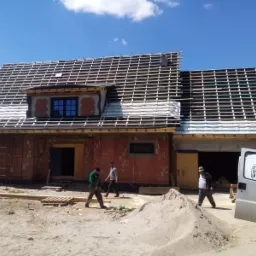 Budowa domu jednorodzinnego z czerwonej cegły, widoczna konstrukcja dachu z łatami i folią, robotnicy na placu budowy, biały samochód dostawczy z otwartymi drzwiami.