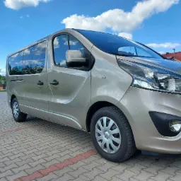 Beżowy van na kostce brukowej, błękitne niebo z chmurami w tle. Auto z przyciemnianymi szybami, gotowe do transportu osób. Widok z boku, lekki skos.