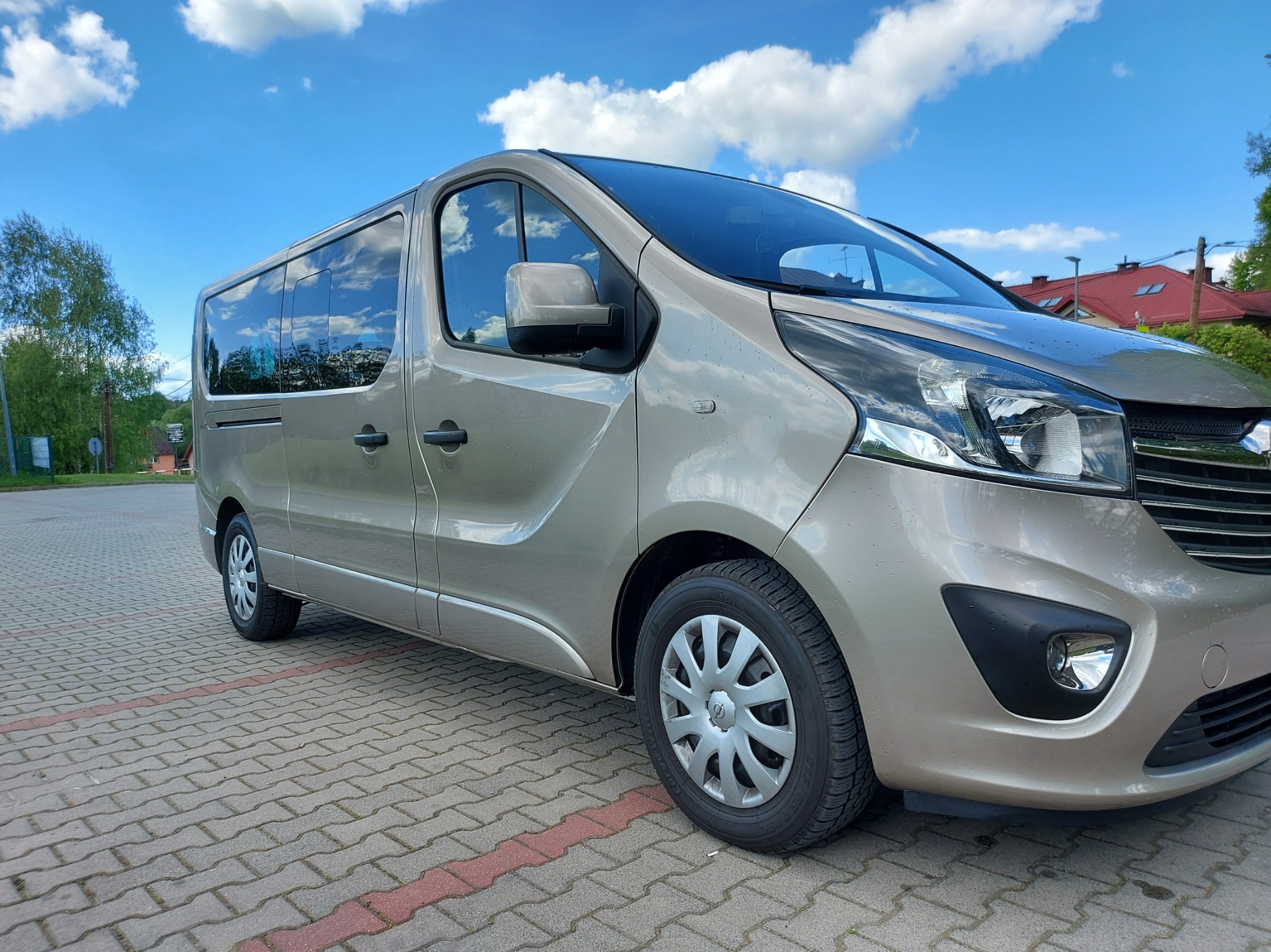 Beżowy van na kostce brukowej, błękitne niebo z chmurami w tle. Auto z przyciemnianymi szybami, gotowe do transportu osób. Widok z boku, lekki skos.