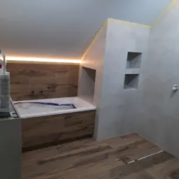 Wykończona łazienka na poddaszu z drewnopodobną podłogą i obudową wanny, podświetleniem LED oraz wnękami w ścianie. Wanna zabezpieczona folią ochronną.