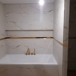 Biała wanna obudowana płytkami imitującymi marmur z złotymi żyłkami, złota armatura, półka w ścianie i złote listwy wykończeniowe.