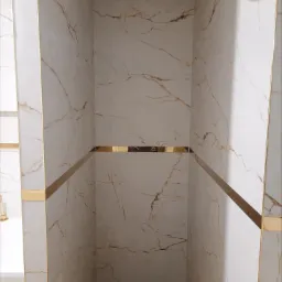 Nowoczesna kabina prysznicowa wyłożona białymi płytkami imitującymi marmur z wyraźnymi złotymi żyłami i dekoracyjnymi złotymi listwami, widoczny odpływ liniowy.