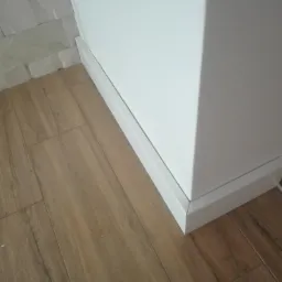 Szczeg&oacute;łowe ujęcie narożnika ściany z białą listwą przypodłogową, panele podłogowe imitujące drewno oraz fragment dekoracyjnej ściany z białego kamienia.